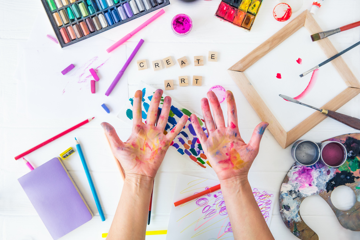 Top view opened painted female hands over Create art words lettering with many colorful paintiing materials on white background. Art workshop, drawing, inspiration, craft, creativity. Art therapy,